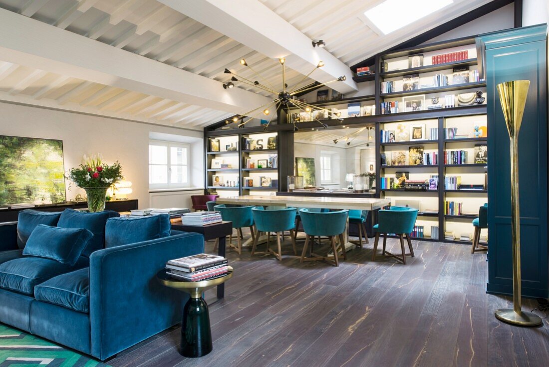 Offener Wohnraum mit blauem Polstersofa, im Hintergrund langer Tisch mit Schalenstühlen vor Regalwand