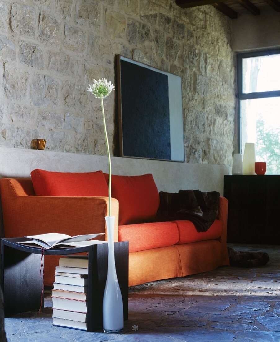 Orange sofa, side table and single allium flower in vase in front of rustic stone wall