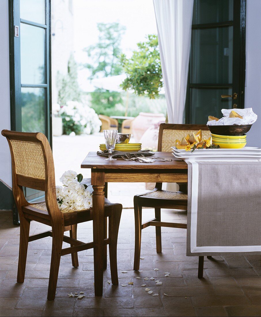 Esstisch mit Geschirr und Besteck vor offener Terrassentür, Geflechtstuhl mit weissen Rosen