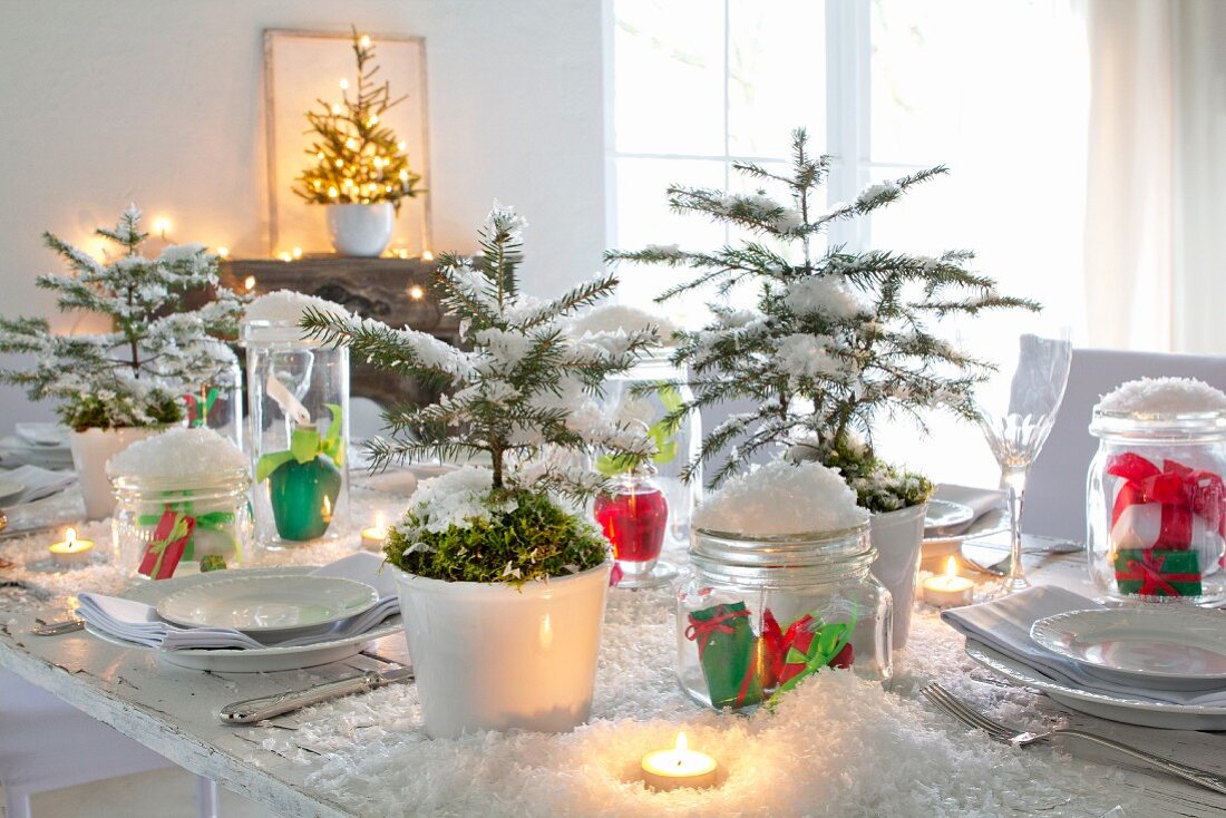 Weihnachtstisch mit Dekoschnee, Fichtenbäumchen, weißem Geschirr und brennenden Teelichtern