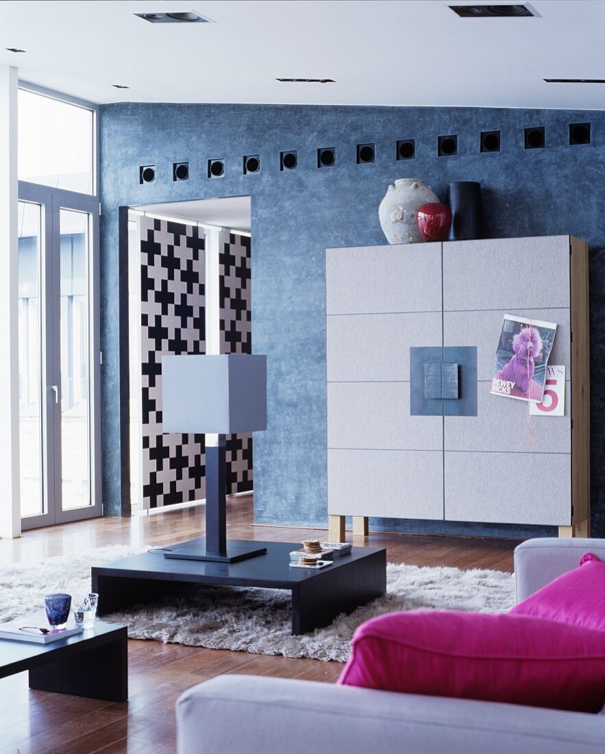 Designer-Schrank mit Filz an blaugrauer Wand davor Flokati mit Bodentisch, Tischleuchte und pinkfarbenen Kissen