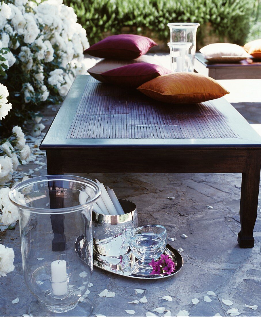 Cushions on coffee table in front of white-flowering rose bushes with candle lantern and silver tray on stone floor