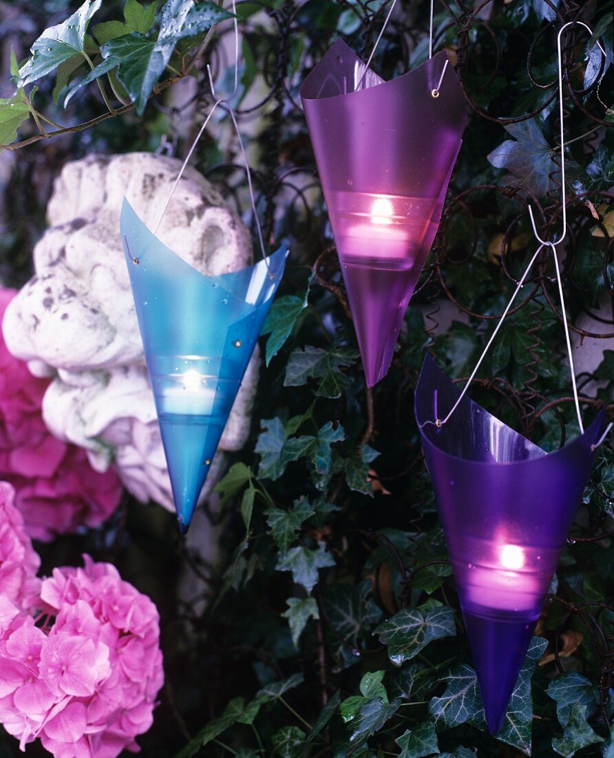 Violette und blaue Windlichthalter in Spitztütenform an efeuberankter Gartenmauer