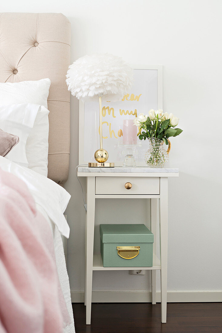 Table lamp with brass base and vase of flowers on white bedside cabinet with brass drawer knob and marble top
