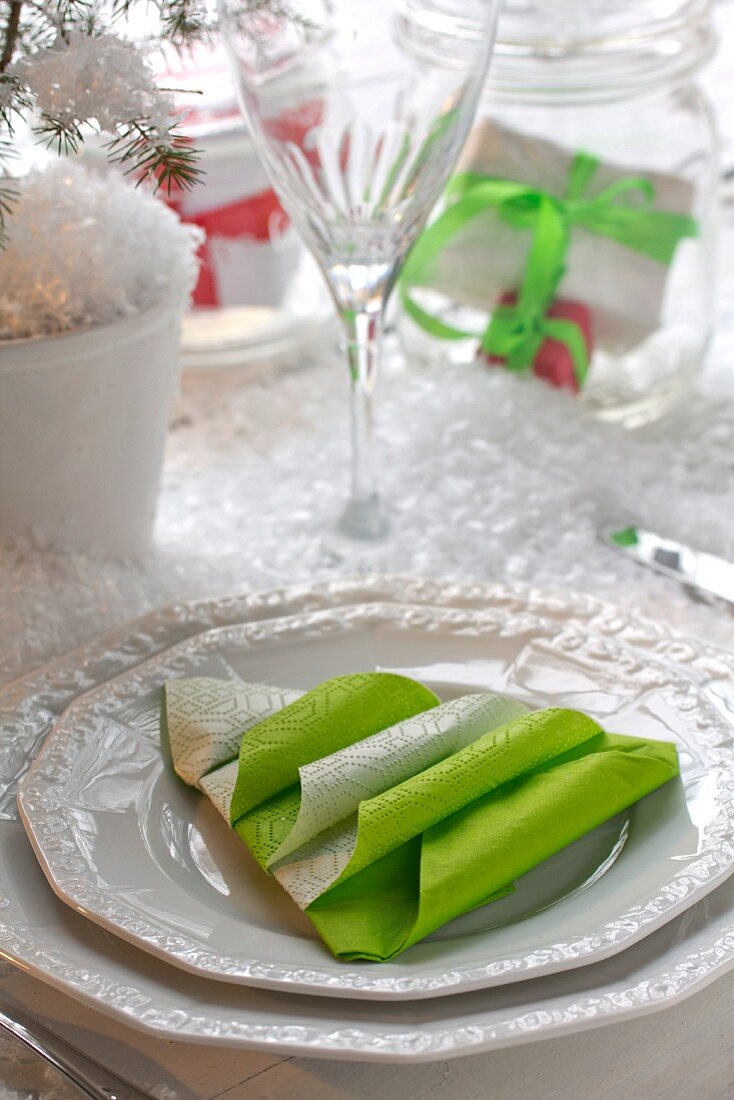 White place setting with green paper napkin folded into Christmas tree amongst artificial snow on table