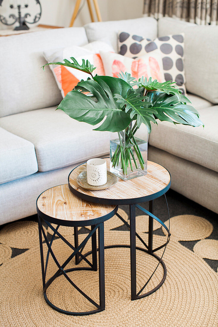 Two-part side table with lantern and philodendron leaves in front of upholstered furniture