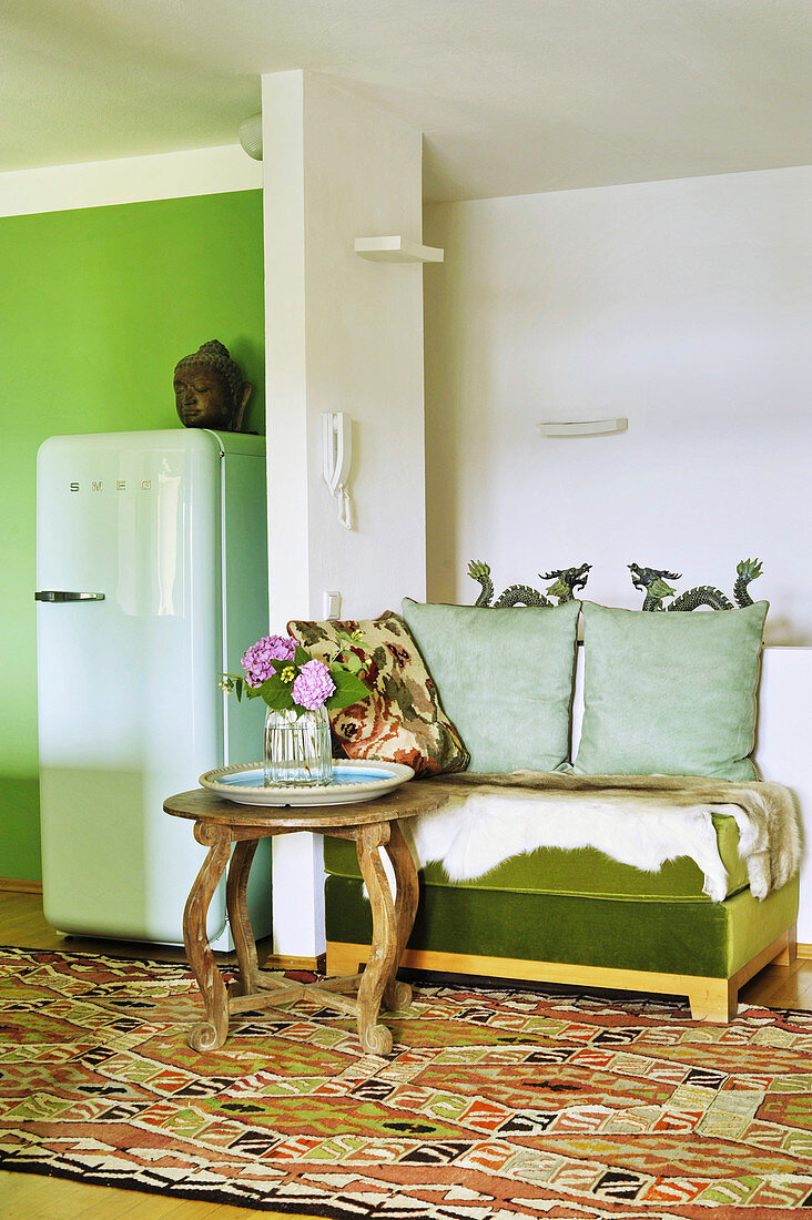 Green bench with animal-skin rug and round table next to partition with fridge on other side