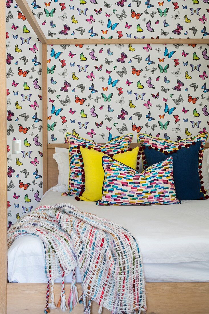 Colourful scatter cushions on modern four-poster bed against butterfly-patterned wallpaper