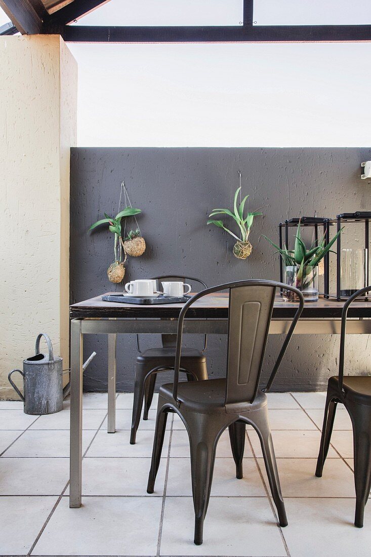 Esstisch mit Metallgestell und Metallstühle auf überdachter Terrasse, hängende Grünpflanzen an grauer Wand