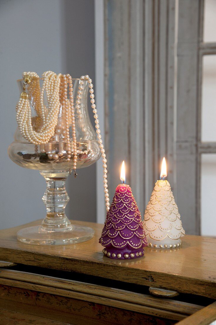 Zwei brennende, weihnachtliche Kerzen neben Glasgefäß mit elegantem Perlenschmuck