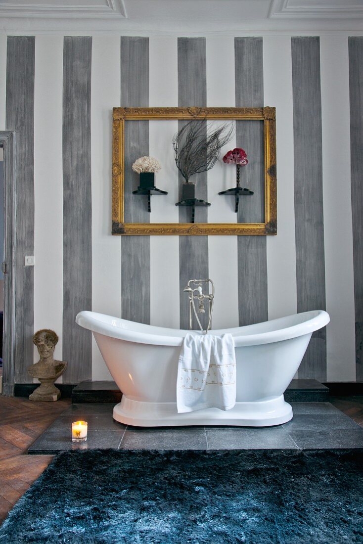 White, free-standing bathtub below gilt picture frame and ornaments on grey-striped wall