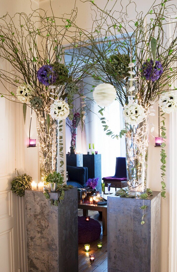 Elegant arrangements of artificial flowers, fairy lights and glass vases on grey plinths in front of living area