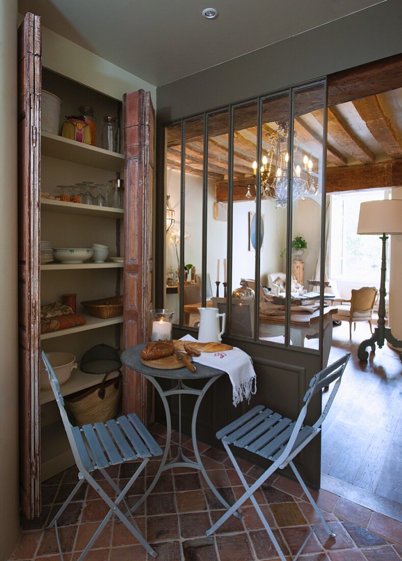 Bistrotisch mit Brot und zwei Klappstühlen vor Geschirrschrank, Blick in Wohnbereich mit Kronleuchter