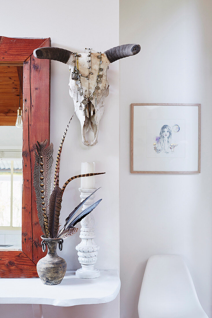 Mirror, vase with feathers and candle holder on white shelf, animal skull above