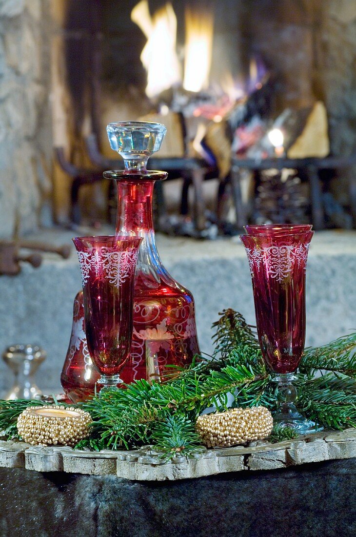 Red carafe and glasses amongst green fir branches in front of open fire
