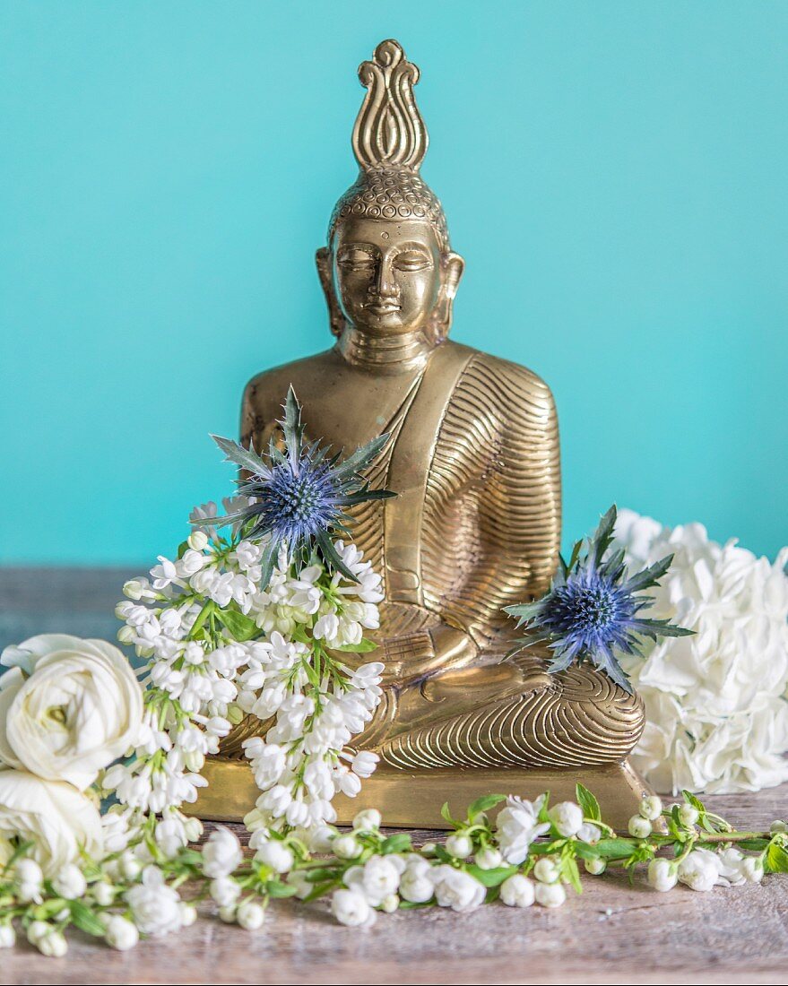 Buddhafigur mit weißen Blüten und Distelblüten vor türkisfarbenem Hintergrund