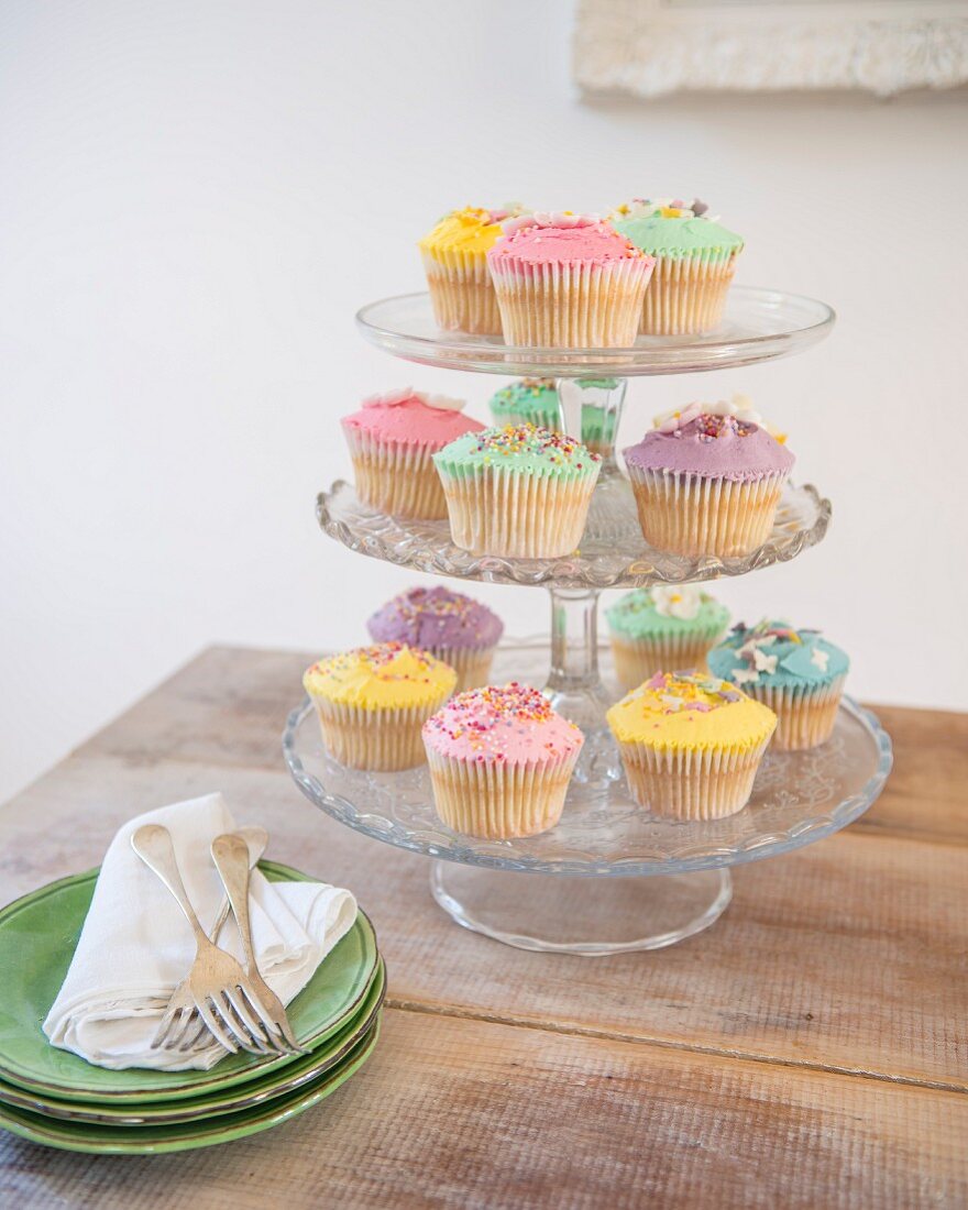 Pastellfarbene Cupcakes mit bunten Zuckerstreusel auf Etagere neben Tellerstapel auf rustikalem Tisch