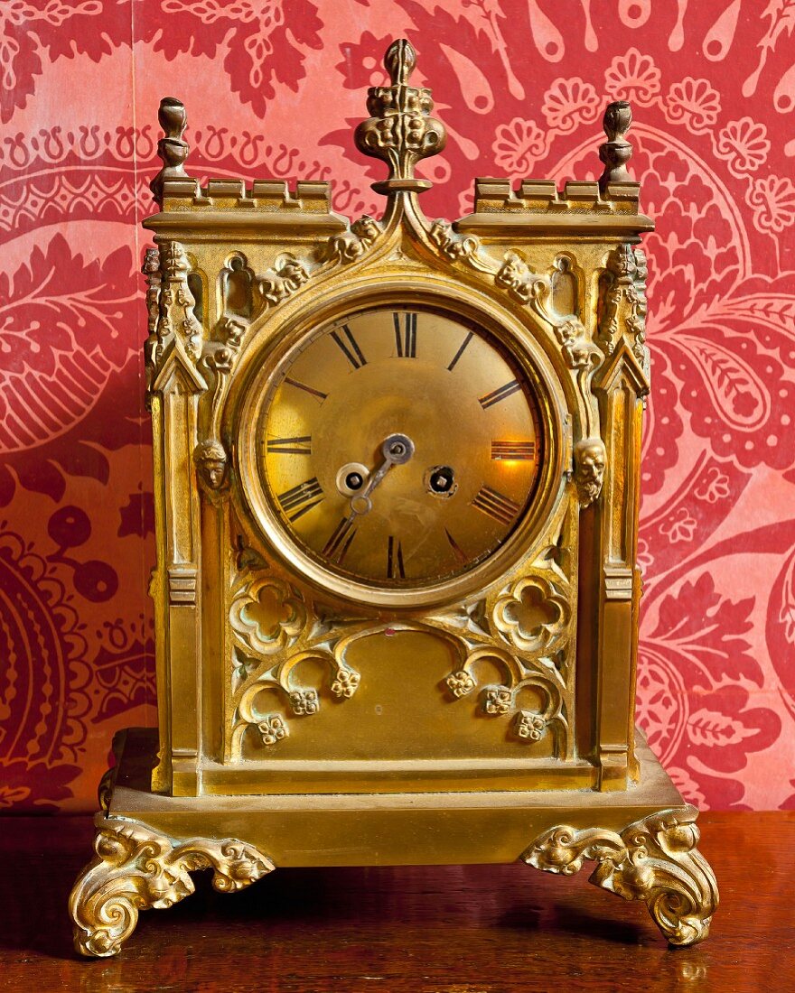 Ornate gilt table clock with Roman numerals against red brocade wallpaper