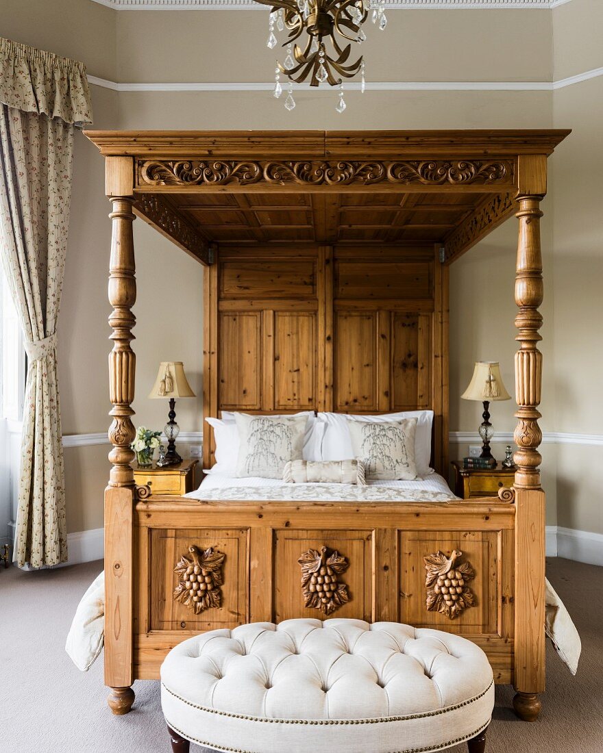 Ornate four-poster bed with cushions in bedroom with bay window and buttoned-tufted stool
