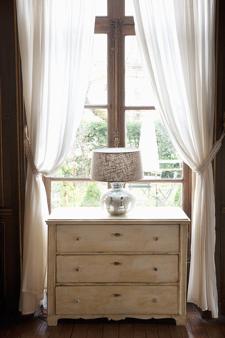 Tischleuchte mit silbernem Fuß auf einer alten Kommode vorm Fenster
