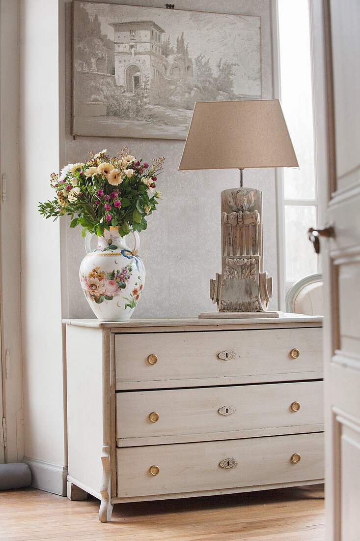 Vase of flowers and table lamp on white chest of drawers seen through open door