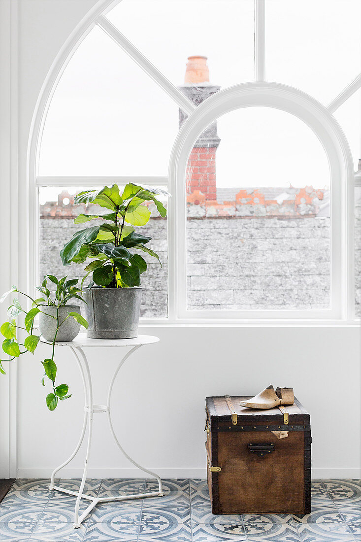 Metalltisch mit Pflanzen und alte Truhe vor einem Bogenfenster