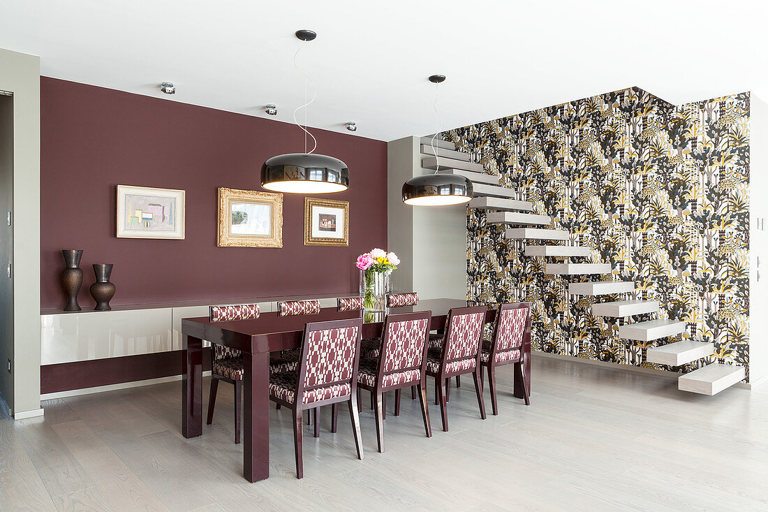 Upholstered chairs in elegant dining area with claret-red wall next to cantilever stairs on wall with patterned wallpaper