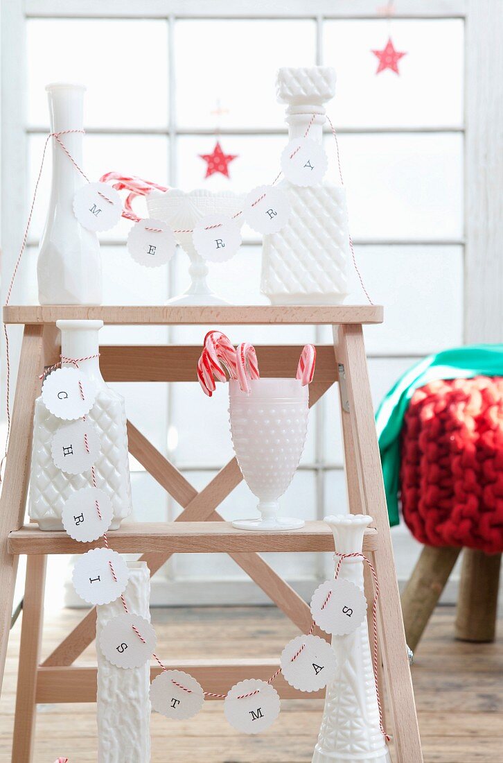 weiße DIY-Papiergirlande mit 'Merry Christmas' und rot-weisser Kordel an weißen Gefäßen