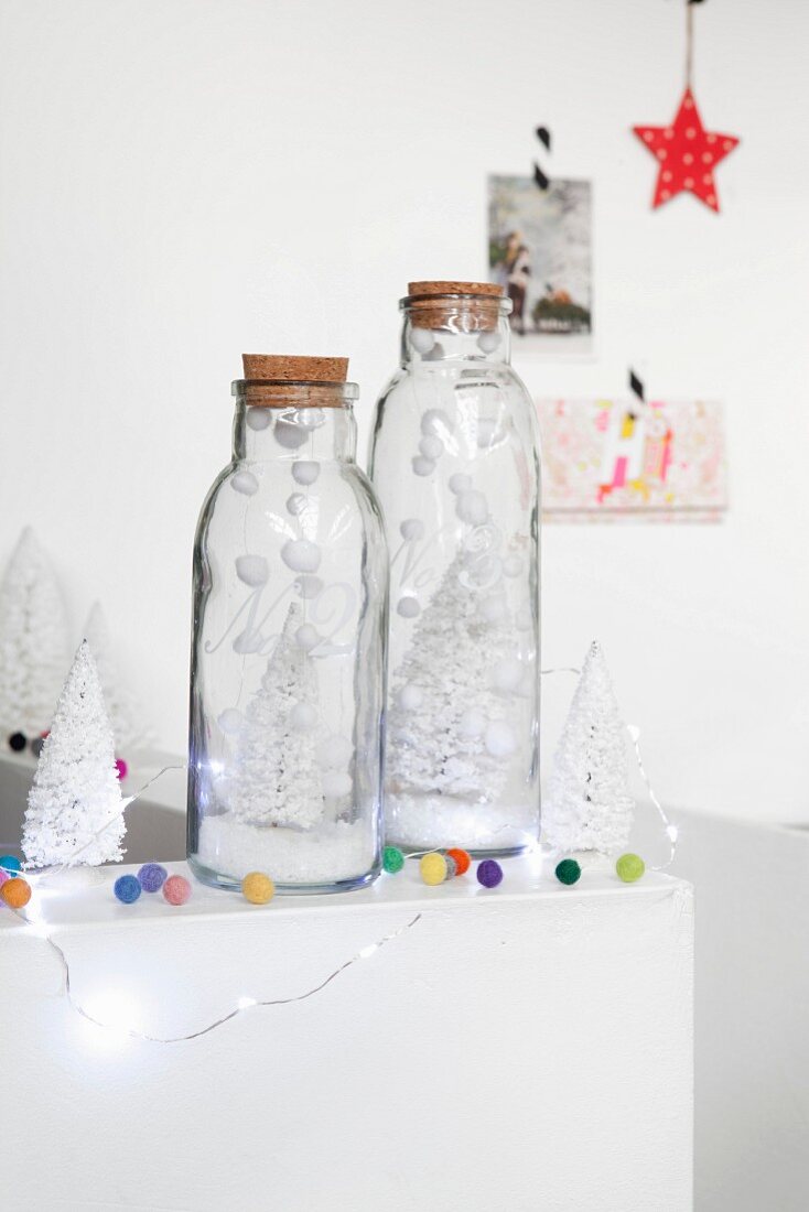 Glasflaschen mit weissen Dekobäumchen, Kunstschnee, bunten Filzkugeln und Lichterkette