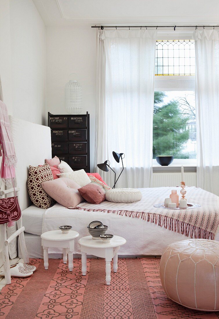 Small white tables, leather pouffe and fringed blanket on bed in feminine bedroom
