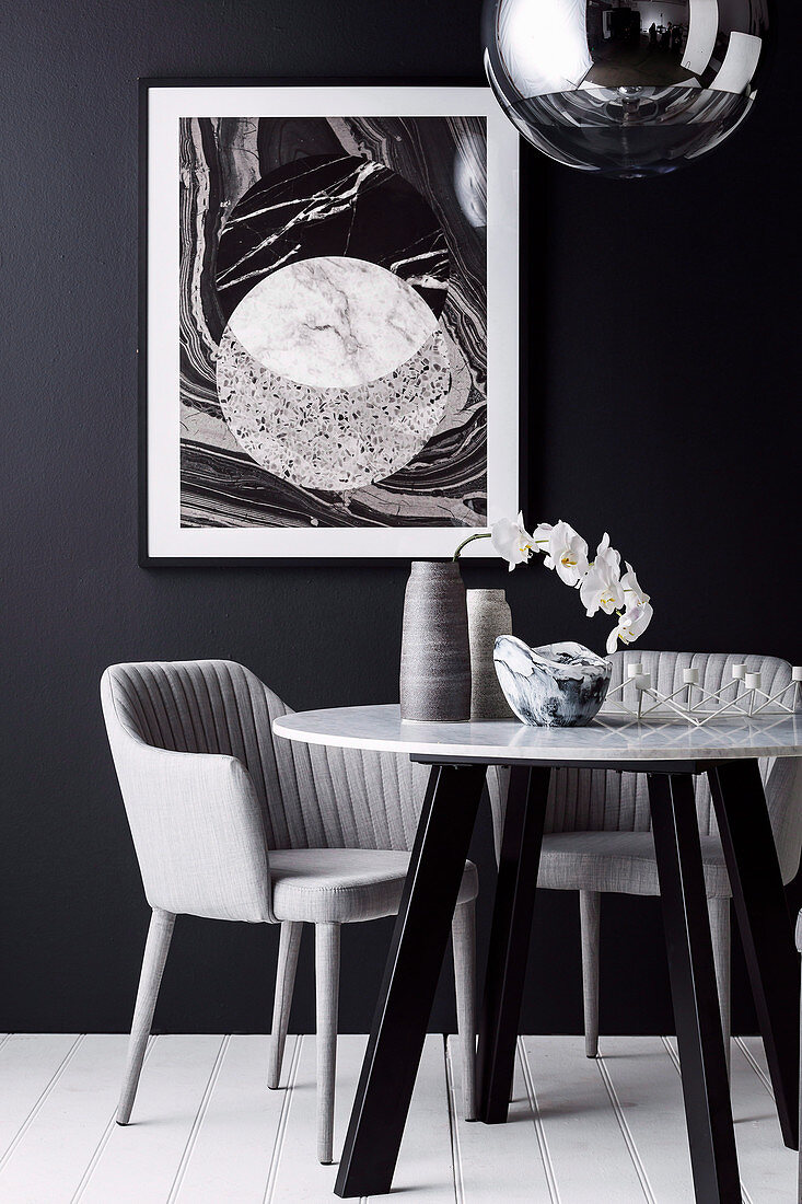 Vase mit Orchidee und Glasschale auf rundem Tisch mit Marmorplatte und Armlehnstuhl vor schwarzer Wand mit Bild