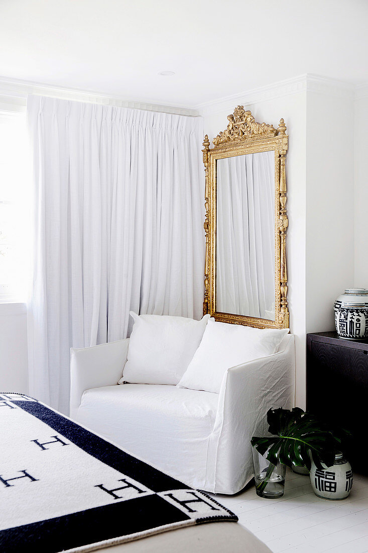 Sessel mit weißer Husse, Goldrahmenspiegel und weißer, bodenlanger Vorhang im Schlafzimmer
