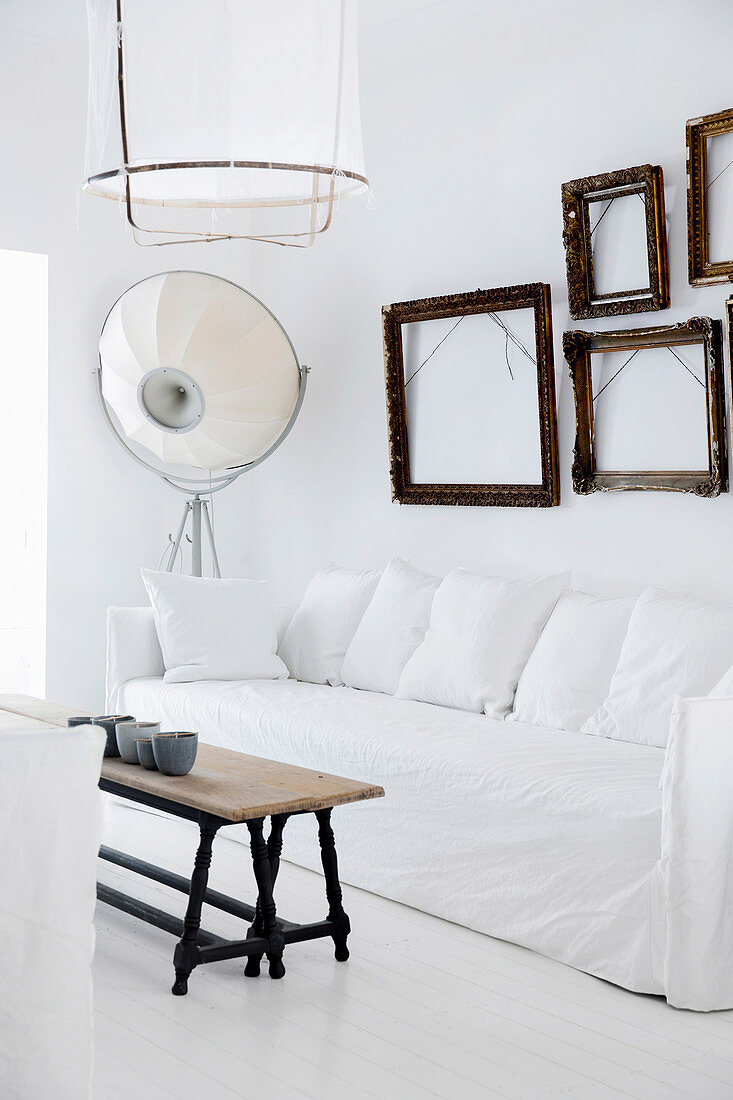Sofa mit weißer Husse und Kissen, darüber leere Bilderrahmen, Vintage Couchtisch auf weißem Dielenboden