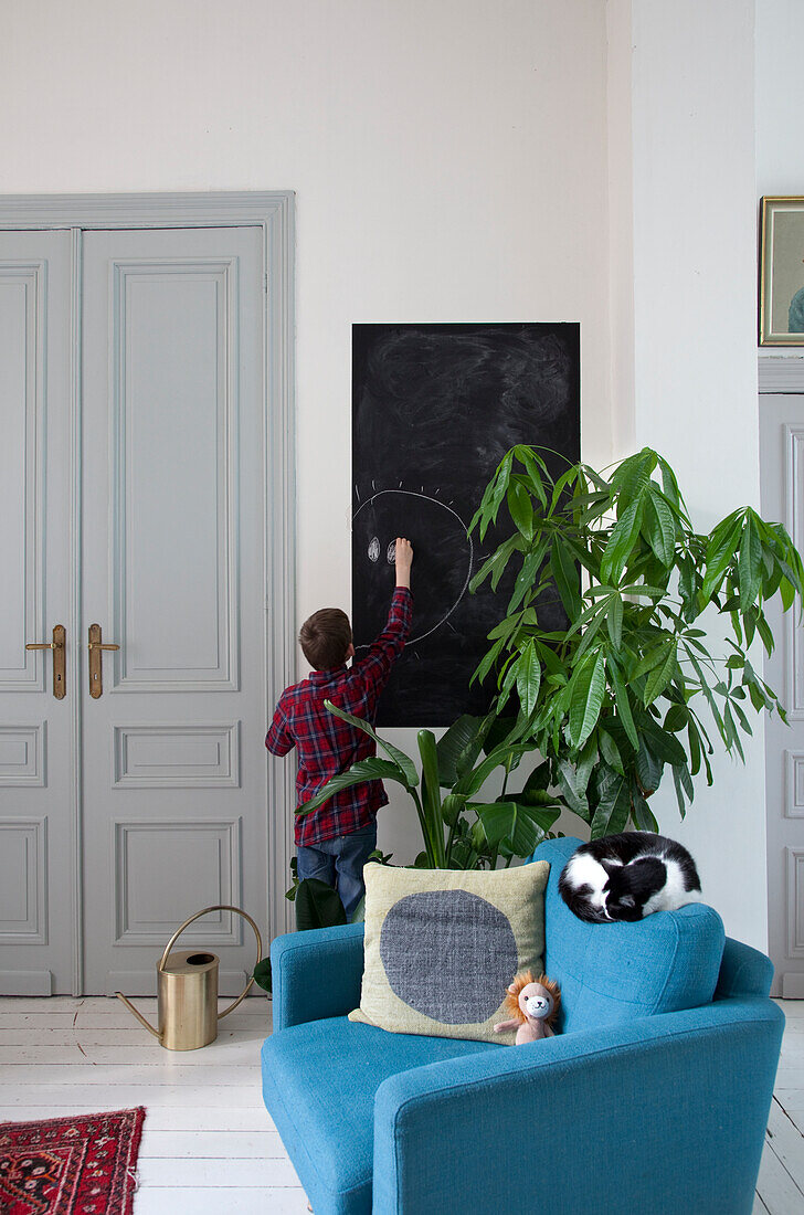 Boy drawing on chalkboard behind blue armchair with cat asleep on backrest