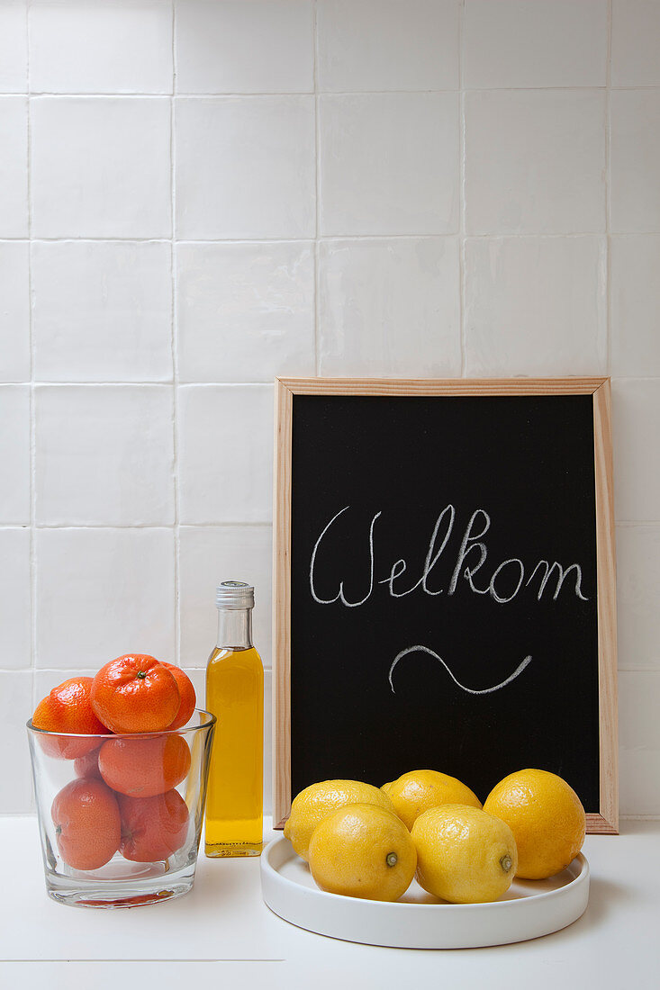 Zitrusfrüchte, Ölflasche und Willkommen-Tafel vor weißer Fliesenwand in der Küche