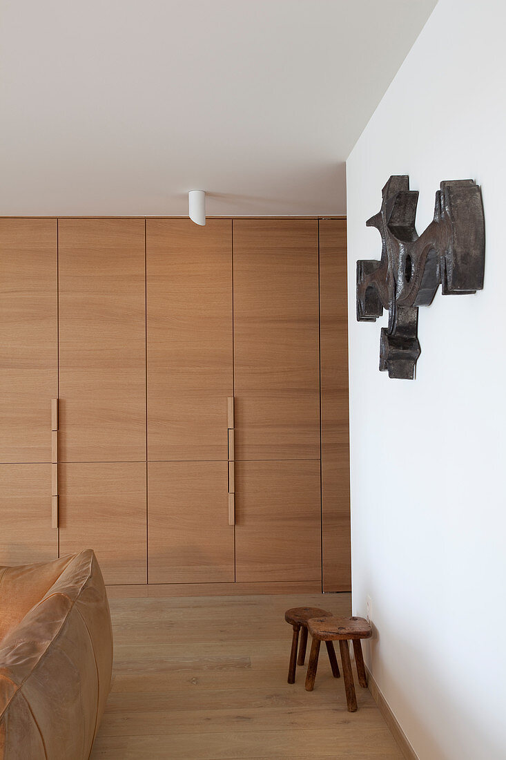 Floor-to-ceiling fitted wardrobes, wooden stools, sculpture on wall and leather couch in living room