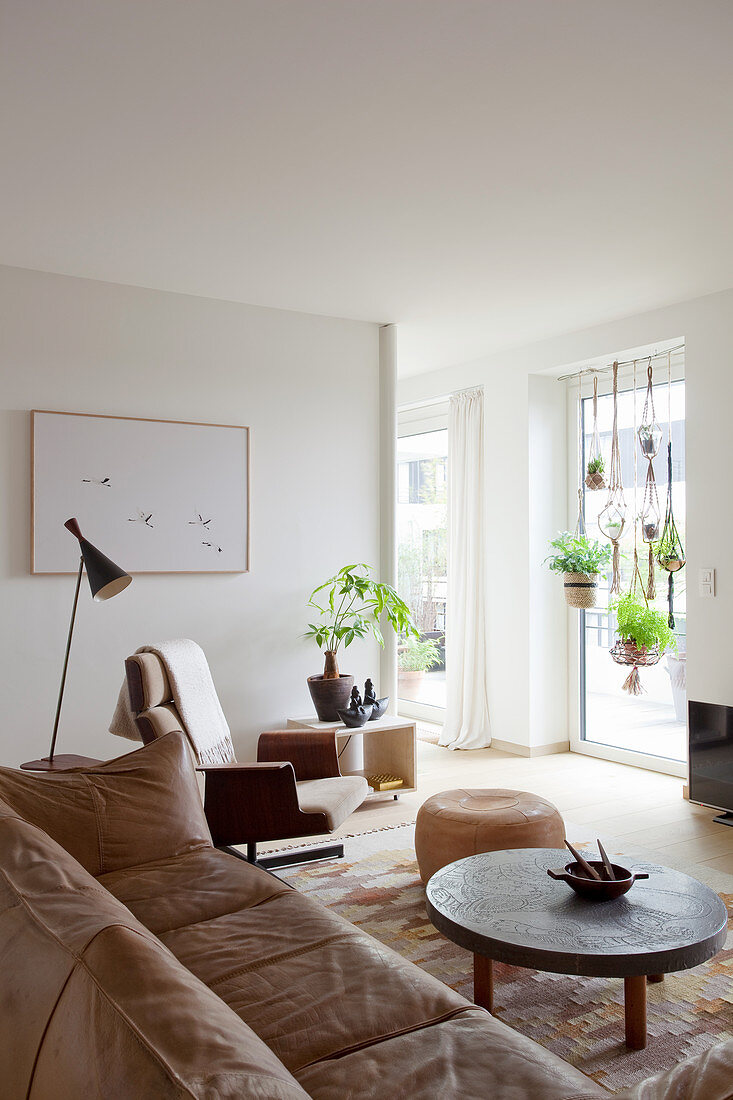 Pale brown couch, coffee table, pouffe and swivel chair in living room with house plants suspended in large window