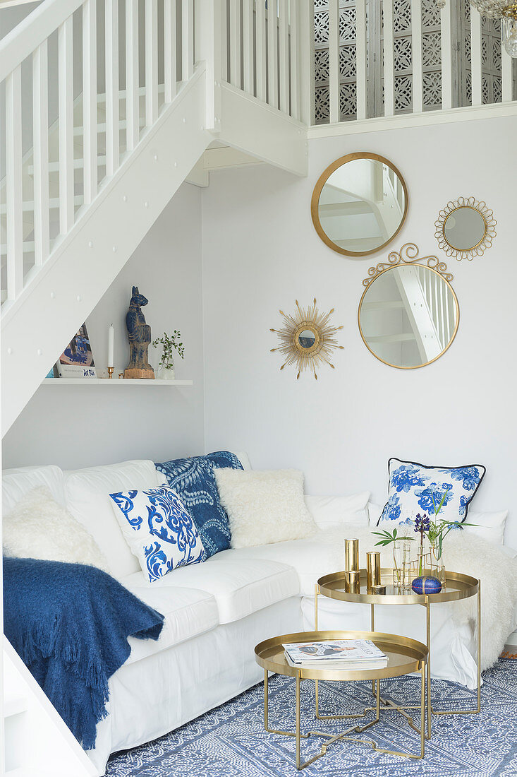 Scatter cushions on white couch and set of round tray tables below staircase with round mirrors on wall