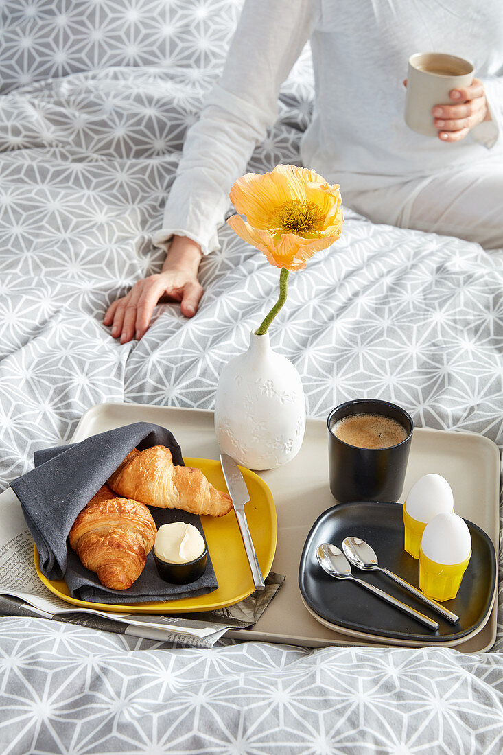 Frühstückstablett mit Croissant, Eiern, Kaffee und gelber Mohnblüte auf dem Bett, Frau im Hintergrund