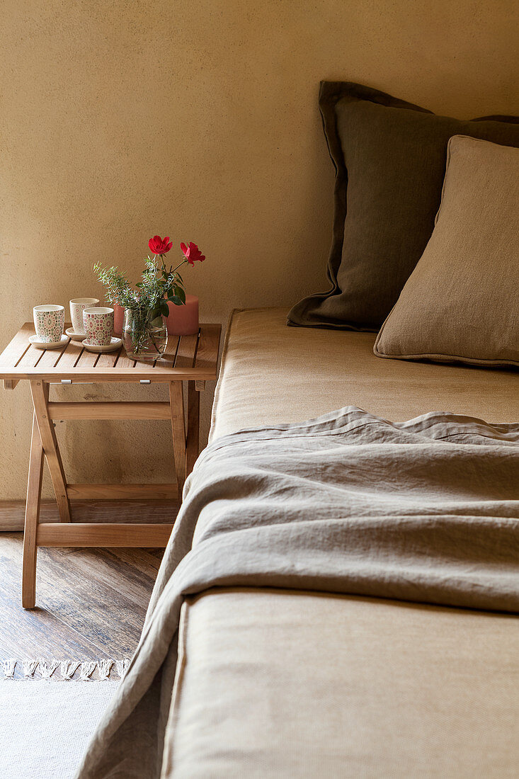 Bett mit Kissen, daneben Holztischchen mit Bechern und Blumen