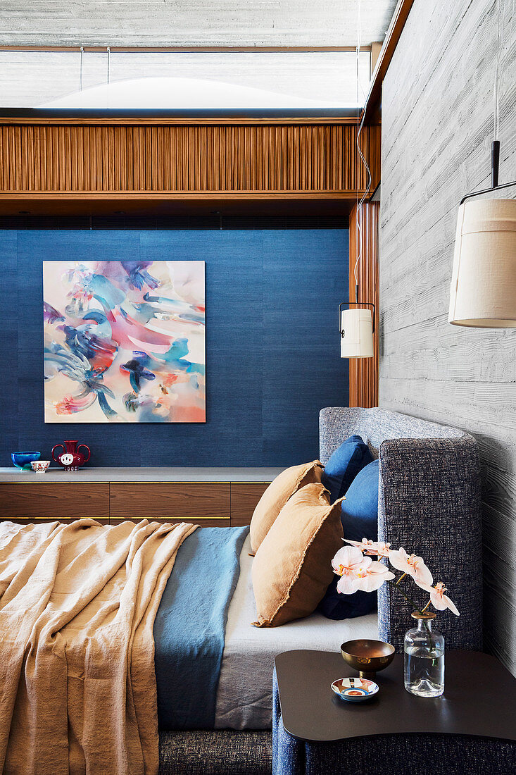 Doppelbett vor Holzwand in Beton-Optik im Schlafzimmer, im Hintergrund Kunstwerk an blauer Wand