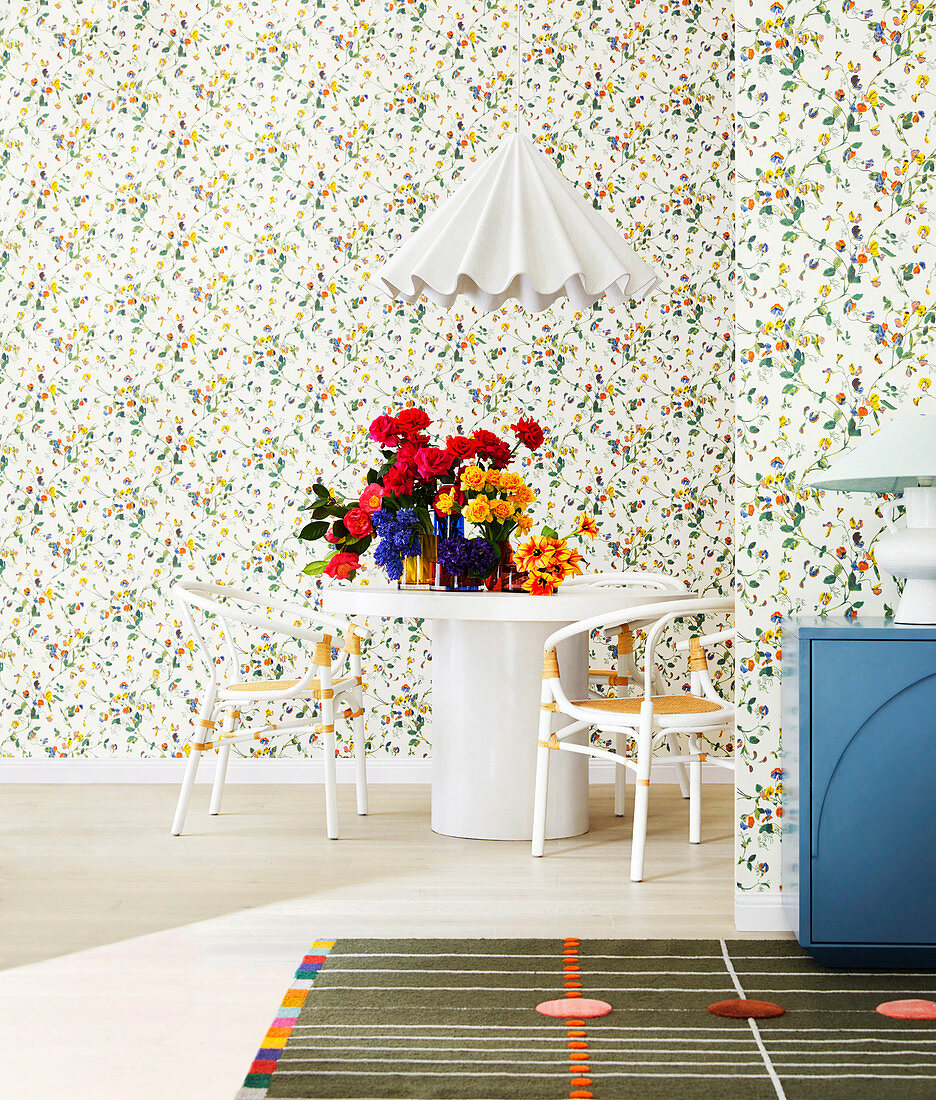 White dining table with flowers and chairs in front of floral wallpaper, white designer hanging lamp