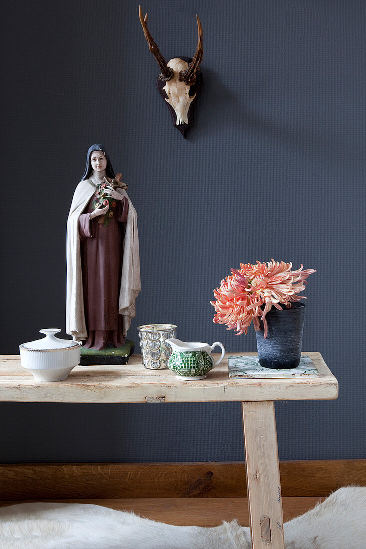 Flowers and Madonna figurine on wooden bench in front of hunting trophy on dark wall