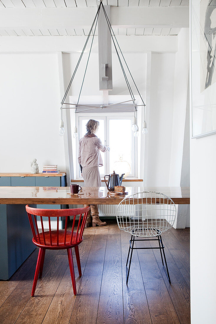 Langer Esstisch mit verschiedenen Stühlen in Wohnküche, im Hintergrund Frau am Fenster