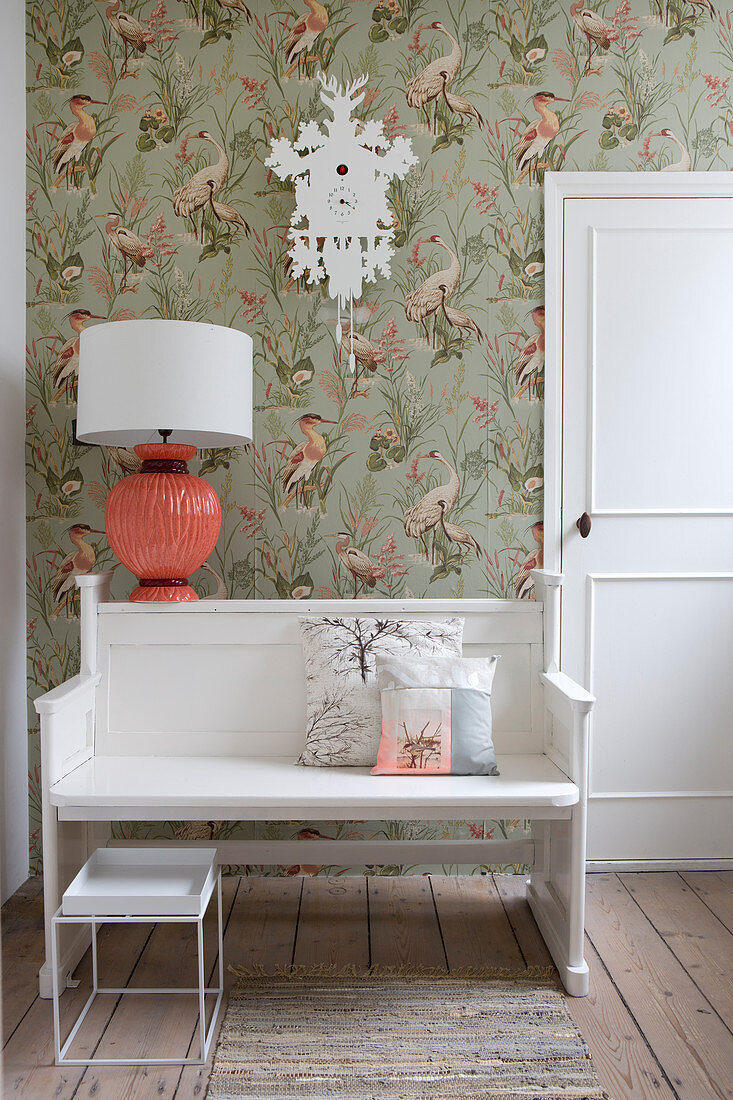 Bench and coral-pink lamp below cuckoo clock on vintage-style wallpaper