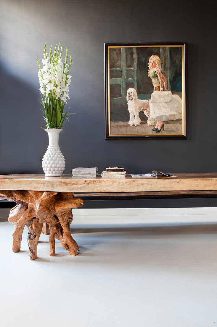 Rustikaler Tisch mit Wurzelholz als Tischbein vor grauer Wand mit Gemälde