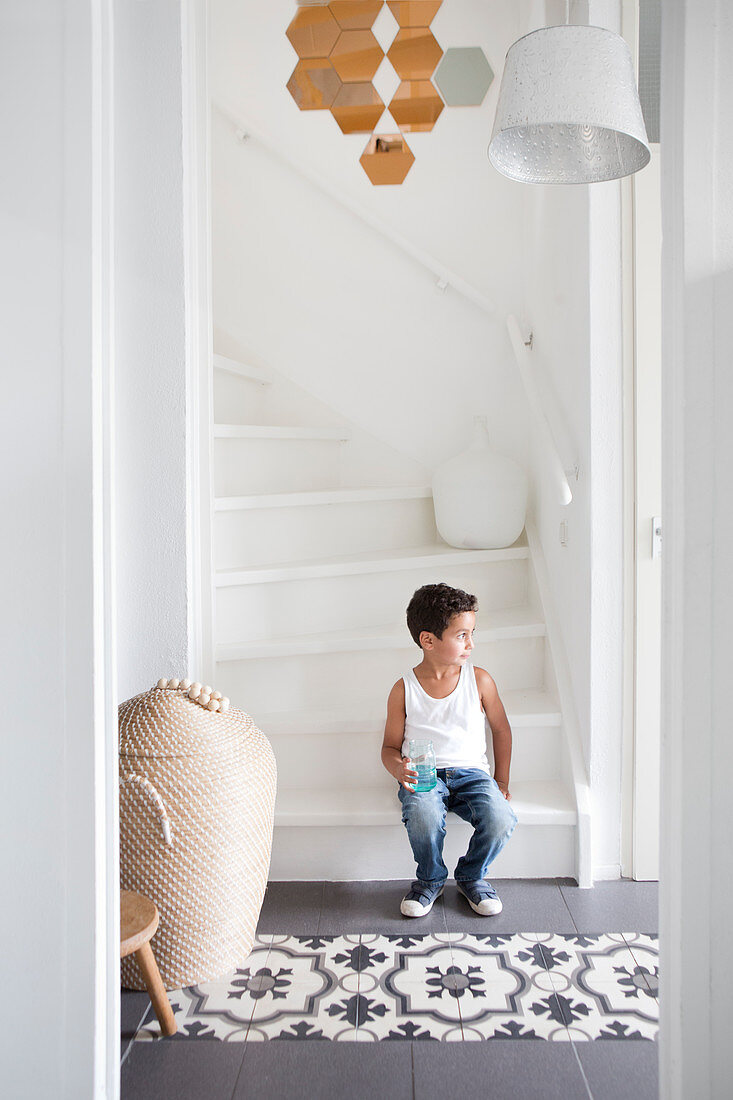 Junge sitzt auf weißer Treppe im Flur mit gemusterten Fliesen