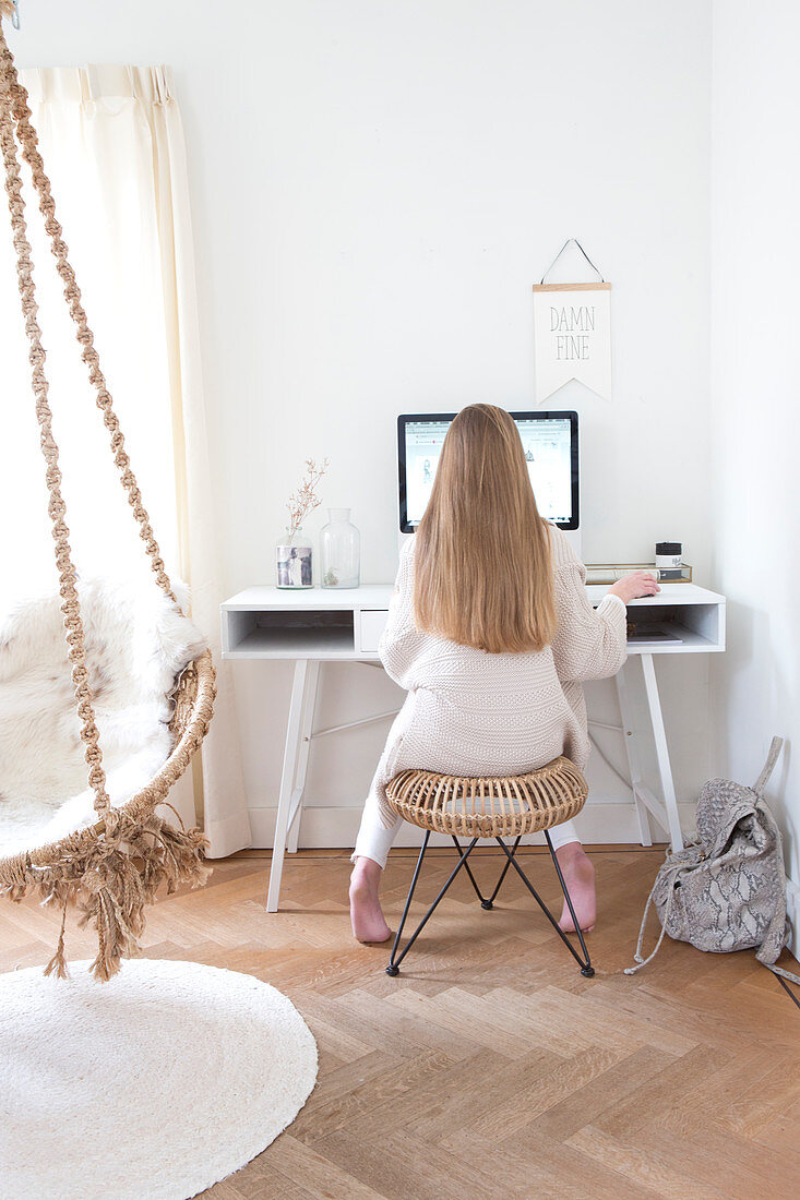 Young woman sitting at a desk in a bright room, in the foreground a macrame hanging chair
