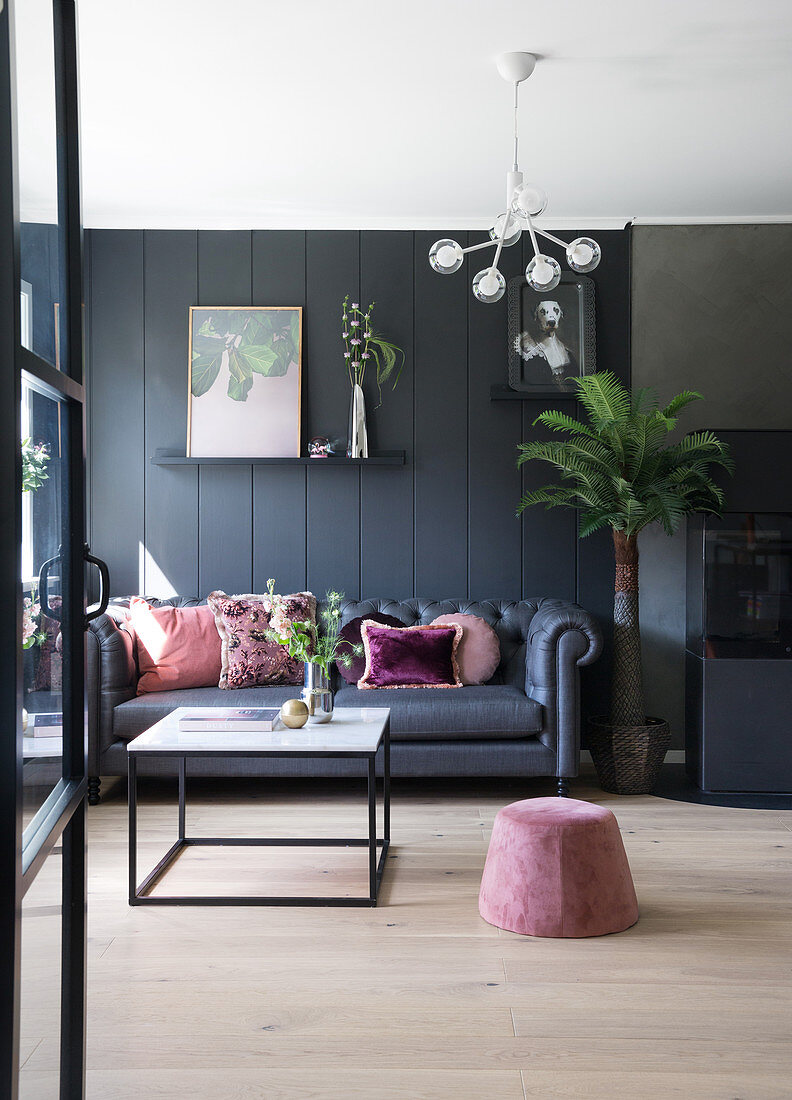 A dark sofa with pink cushions in front of a dark wall with a pouffe and a coffee table in the foreground
