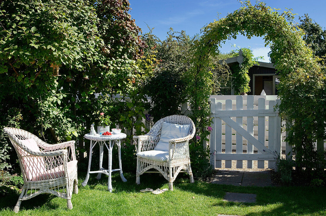 Sitzplatz im Garten mit Korbmöbeln neben dem Gartentörchen unterm Rankbogen